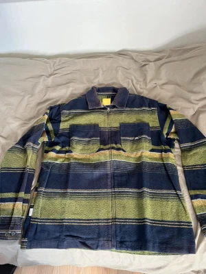 Randig overshirt i blått och grönt - Overshirt med breda horisontella ränder i marinblått, olivgrönt och inslag av gult. Två bröstfickor med lock, dragkedja framtill och klassisk krage. Långärmad och tillverkad i ett mjukt, vävt material som ger en avslappnad look.