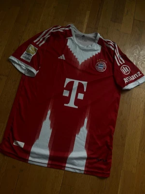 Bayern München matchtröja Musiala - Röd och vit Bayern München matchtröja köpt i Bosnien, med Musiala och nummer 42 på ryggen. Tröjan har klubbmärke, sponsorer och vita detaljer på ärmar och axlar. Tillverkad i lätt och ventilerande material, perfekt för fotbollsträning eller match.