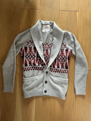 Grå cardigan från Hollister - Säljer en grå kofta från Hollister med sjalkrage och knäppning framtill. Koftan har ett vinrött och vitt geometriskt mönster över bröstet och två fickor fram. Perfekt för dig som gillar en avslappnad men snygg stil.