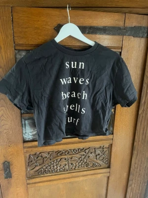 Svart croppad t-shirt KappAhl - Snygg svart croppad t-shirt från KappAhl med vit text på framsidan. T-shirten har korta ärmar och rund halsringning. Perfekt för dig som gillar en avslappnad och trendig stil.