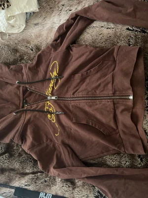 Brun Ed Hardy hoodie med tryck - Säljer en brun hoodie från Ed Hardy med dragkedja, huva och snörning. Framsidan har guldfärgat Ed Hardy-tryck och fickor, medan baksidan har ett stort färgglatt tatueringsinspirerat motiv med hjärta och blommor. Perfekt för dig som gillar streetwear och unika detaljer.