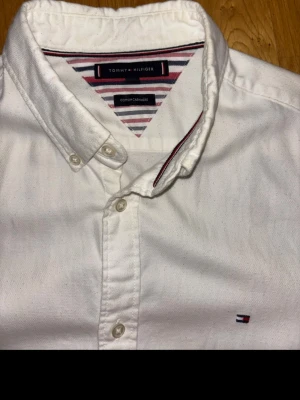 Vit cashmere skjorta från Tommy Hilfiger - Klassisk vit skjorta från Tommy Hilfiger med knappslå och diskret logga broderad på bröstet. Skjortan har långa ärmar, button down-krage och är tillverkad i bomull. Snygga randiga detaljer på insidan av kragen ger extra stilpoäng. ”Diffrent guys, they rockin’ Polo, I’m rockin’ Tommy Hil” - Lil Durk.