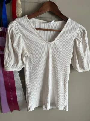 Vit ribbad topp med puffärm - Snygg vit ribbad topp med puffiga kortärmar och v-ringning. Tunn och stretchig, perfekt till jeans eller kjol. Enkel men trendig design som passar till många olika stilar.