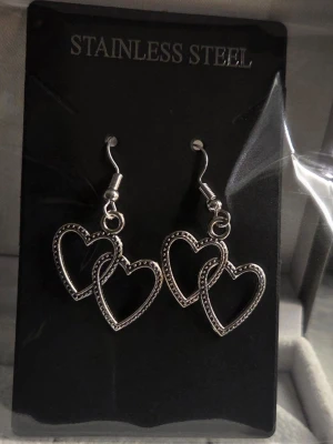 Silverfärgade hjärtörhängen i stål - Snygga örhängen i silverfärgat rostfritt stål med dubbla hjärtan som hänger. Hjärtana har små detaljer längs kanterna som ger en cool touch. Perfekta för dig som gillar statement-smycken och vill ha något unikt till din stil.
