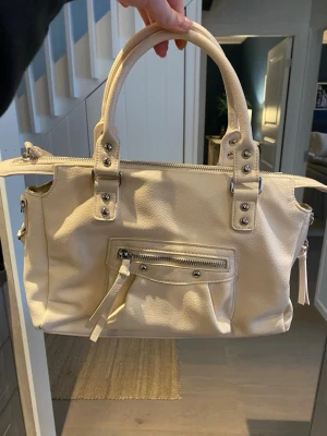 Krämvit handväska med nitar - Snygg krämvit handväska i mjukt konstläder med silverfärgade nitar och detaljer. Väskan har två rejäla handtag, en dragkedjeficka framtill och dragkedja upptill. Perfekt storlek för det viktigaste och ger en clean look till din outfit.
