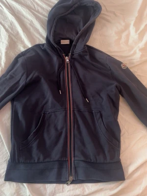 Mörkblå hoodie från Moncler ÄKTA Storlek S - Snygg mörkblå hoodie från Moncler med dragkedja framtill, två stora fickor och justerbar huva med snören. Klassisk Moncler-logga på ärmen och ribbade muddar. Tillverkad i mjuk bomull för skön känsla och stilren look.