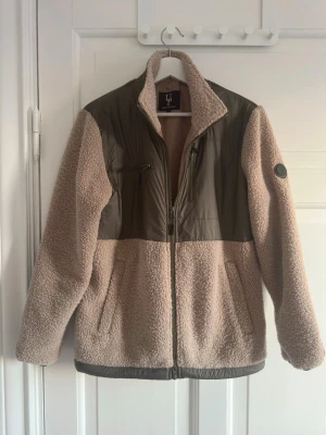 Vår jacka från  - Teddyjacka i beige med kontrasterande gröna partier på axlar, bröst och ärmar. Dragkedja framtill, två sidofickor och en bröstficka med dragkedja. Mjuk, fluffig teddyfleece och hög krage. 