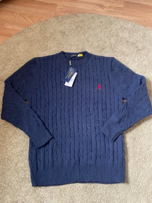 Blå kabelstickad tröja Polo Ralph Lauren - Mörkblå kabelstickad tröja från Polo Ralph Lauren med rund halsringning och röd broderad logga på bröstet. Tröjan har ribbade muddar vid ärmslut och nederkant. Perfekt för dig som gillar klassisk och stilren design.