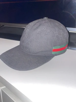 Svart Gucci keps med GG-mönster - Säljer en svart keps från Gucci med det klassiska GG-monogrammet över hela kepsen. På sidan finns en grön och röd rand som ger den en ikonisk touch. Kepsen har böjd skärm och är tillverkad i ett slitstarkt tygmaterial. Perfekt för alla årstider , priset är inte hugget i sten 