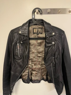 Svart skinnjacka från Jofama - Snygg svart skinnjacka från Jofama by Marie Serneholt. Klassisk bikerstil med silvriga dragkedjor, bred krage och dekorativa sömmar. Fodret har ett diskret mönster i grått och rött. Perfekt för dig som vill ha en tidlös och cool look.