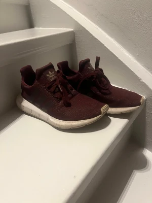 Vinröda Adidas sneakers - Snygga vinröda Adidas sneakers med klassiska tre ränder på sidan och vit sula. Ovandelen är i textilmaterial och skorna har snörning. Perfekta för dig som vill ha en stilren och sportig look.