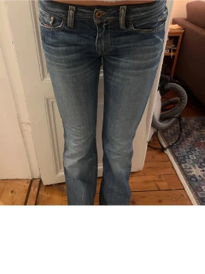 Blå bootcut jeans från diesel - Säljer ett par blå bootcut low wast jeans från Disa med klassisk femficksdesign och snygga slitningar. Jeansen har broderade detaljer på bakfickorna och en bekväm passform som sitter snyggt över höften. Perfekt för dig som gillar en tidlös och cool look.
