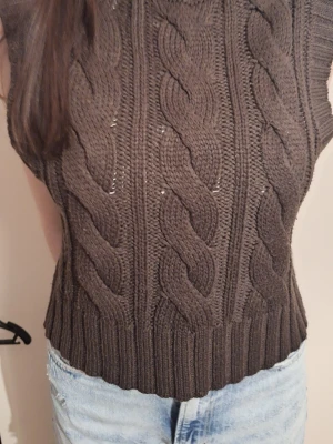 Brun stickad kabelväst med polokrage - Snygg brun stickad väst med grovt kabelstickat mönster och hög polokrage. Perfekt att styla över en skjorta eller t-shirt för en trendig lager-på-lager-look. Västen har ribbade kanter och är ärmlös för en modern vibe.