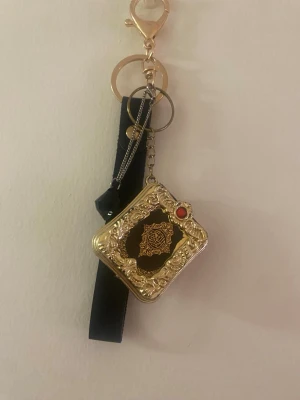 Dekorativ nyckelring med gulddetaljer - Säljer en unik nyckelring med fyrkantig berlock i guld och svart, dekorerad med orientaliskt mönster och en röd sten. Nyckelringen har även en svart banddetalj och en svart kristall. Perfekt accessoar för dig som gillar detaljer och vill sticka ut.