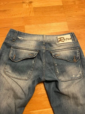 Blå Retro jeans med stora bakfickor - Säljer ett par blå jeans från Retro med slitna detaljer och stora fickor bak med lock. Jeansen har en rak passform och ljusa tvättade partier för en cool vintagekänsla. Klassisk femficksmodell med bälteshällor och knappgylf.