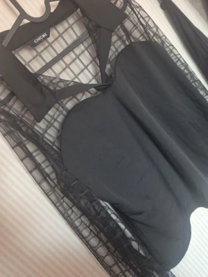 Svart meshblus med krage från Chique - Trendig svart blus från Chique med transparenta meshärmar i rutigt mönster och klassisk krage. Kroppen är i solid svart tyg och ärmarna är långa och genomskinliga, vilket ger en cool kontrast. Perfekt för dig som vill sticka ut med en edgy look.