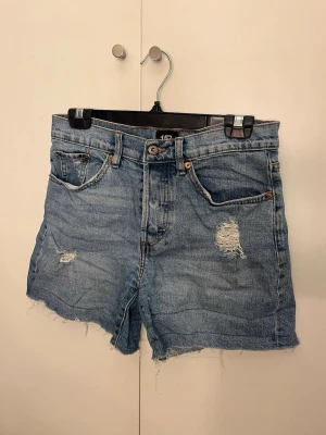 Blå denimshorts med slitningar - Säljer ett par blå denimshorts med råa kanter och slitna detaljer framtill. Klassisk femficksmodell med knappgylf och bälteshällor. Perfekta för en avslappnad och trendig sommarlook. Aldrig använda
