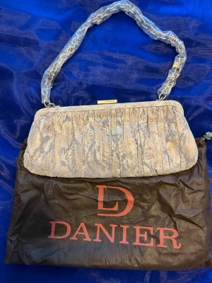 Beige kuvertväska från Danier - Snygg beige kuvertväska från Danier med ormmönstrad yta och guldfärgad metallknäppning. Väskan har en kedjerem insvept i plast och ett ljust innerfoder med dragkedjefack. Perfekt för dig som gillar stilrena detaljer och trendiga accessoarer.