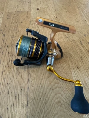 Daiwa fiskerulle i guld och svart - Säljer en Daiwa fiskerulle i guld och svart med detaljer i silver och svart. Handtaget har ett ergonomiskt grepp i svart och guldfärgad vev. Rullen är tillverkad i metall och plast och har en modern design, perfekt för fiskeentusiasten.