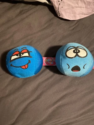 Blå mjuka stressbollar med ansikten - Två blå stressbollar i tyg med tryckta ansikten, en med busigt uttryck och en med förvånad min. Perfekta att klämma på när du behöver slappna av eller vill ha något kul på skrivbordet. Säljs ihop!