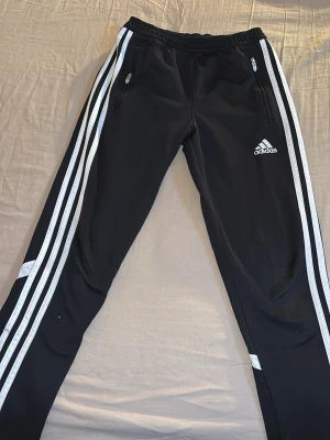 Svarta Adidas mjukisbyxor med vita ränder - Säljer ett par svarta Adidas mjukisbyxor med klassiska vita ränder längs benen och dragkedjeförsedda fickor. Byxorna har Adidas-logga på låret och är tillverkade i ett lätt och ventilerande material (Climacool). Perfekta för chill eller träning.