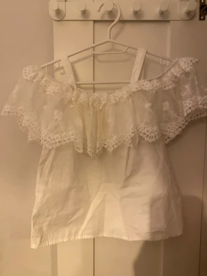 Vit offshoulder topp med spetsvolang - Söt vit offshoulder topp med bred spetsvolang runt axlarna och axelband. Toppen har romantisk känsla och är perfekt för varma dagar. Tillverkad i bomull med fina spetsdetaljer som ger en extra touch.