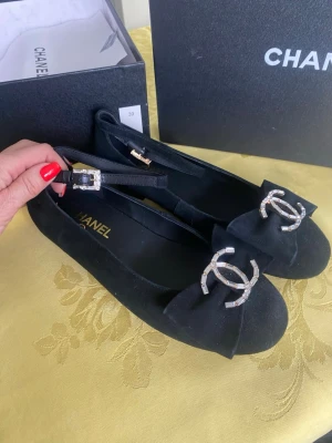 Svarta ballerinaskor från Chanel 38/39 nya med Swarovski  - Stilrena svarta ballerinaskor från Chanel i mocka med dekorativt CC-logo i strass framtill. Skorna har en elegant rem med glittrande spänne och rundad tå. Tillverkade i Italien och levereras med originalkartong.