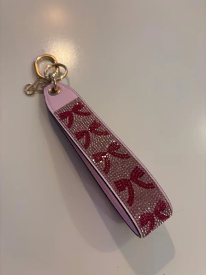 Rosa nyckelband med glitter och rosetter - Säljer ett rosa nyckelband med glittriga detaljer och röda rosetter. Bandet har en bred form och är dekorerat med små kristaller. Metallringen är guldfärgad och bandet är gjort av konstläder och plast. Perfekt för att hålla nycklar eller som accessoar.
