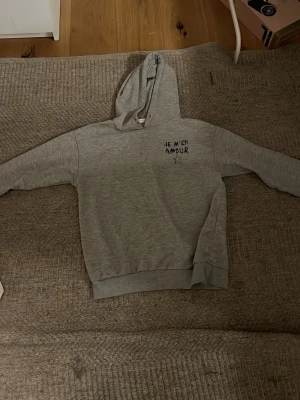 Grå hoodie med Je M'en Amour tryck - Grå hoodie med huva och svart tryck på bröstet där det står 'JE M'EN AMOUR' samt en stjärna under texten. Hoodien har ribbade muddar vid ärmslut och nederkant. Perfekt för en avslappnad och cool look.