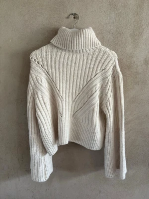 Vit ribbstickad polotröja - Säljer en krämvit ribbstickad polotröja med breda ärmar och höga muddar. Tröjan har ett snyggt mönster framtill och en hög, tjock polokrage som ger en trendig look. Perfekt för lager på lager och riktigt mysig under kalla dagar.