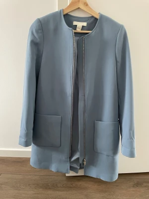 Ljusblå kappa H&M - Stilren ljusblå kappa från H&M med rak passform, rund halsringning och två stora fickor framtill. Dragkedja hela vägen och släta ärmar ger en clean look. 