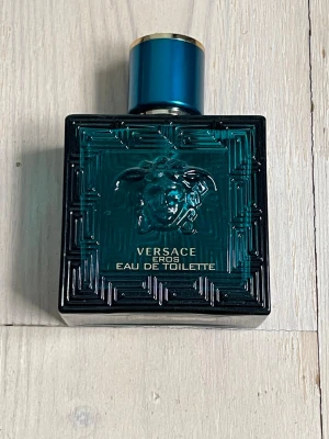 Versace Eros Eau de Toilette - Versace Eros Eau de Toilette i en stilren, fyrkantig flaska av mörk turkost glas med Medusa-huvudet i relief och gulddetaljer. Flaskan har en lyxig känsla och lock med guldfärgad Medusa-symbol på toppen. Perfekt för dig som gillar exklusiva dofter. Köpt på åhlens i mall of dcsnfinavia förra året. OBS Saknar deoderanten få jag tappade bort den. Ca halvfull.
