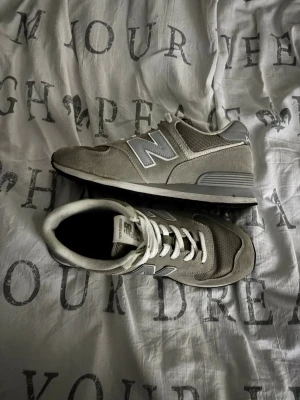 Grå New Balance sneakers - Snygga grå sneakers från New Balance med klassisk N-logga på sidan. Skorna har en kombination av mocka och mesh, vita skosnören och en vit/grå sula. Perfekta för en avslappnad streetstyle-look.