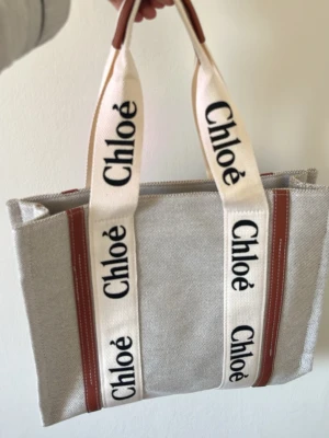 Chloé Woody beige handväska - Trendig beige handväska från Chloé med breda vita handtag där loggan syns tydligt i svart. Väskan är tillverkad i canvas med bruna detaljer i läder och har ett rymligt fack som rymmer laptop och andra essentials. Perfekt för dig som gillar stilren design och logotypdetaljer.