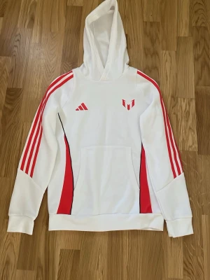 Vit och röd hoodie från Adidas - Snygg vit hoodie från Adidas med röda detaljer och klassiska tre ränder på ärmarna. Hoodien har en stor ficka framtill och huva. Adidas-logga och ett rött emblem på bröstet. Perfekt för en sportig och avslappnad stil. Tröjan är Messi loga hans egna märke är helt oanvänd och inget slitage.