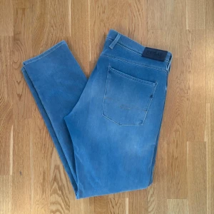 Replay Anbass - Snygga ljusblå slim fit jeans från Replay med klassisk femficksdesign och diskreta slitningar framtill. Jeansen har normal midja, bälteshällor och är tillverkade i mjukt denimtyg som ger en skön känsla. Perfekta för en avslappnad och trendig look. Små vita fläckar vid fickan men inget speciellt.
