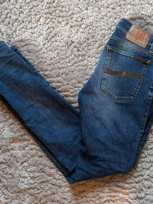 Nudie Jeans W28 L32 - Säljer ett par klassiska blå jeans från Nudie Jeans med fem fickor och snygga slitningar framtill. Jeansen har en smal passform och är gjorda i mjukt denimtyg med Nudie-loggan på bakfickan. Perfekta för dig som gillar stilrena och tidlösa jeans.