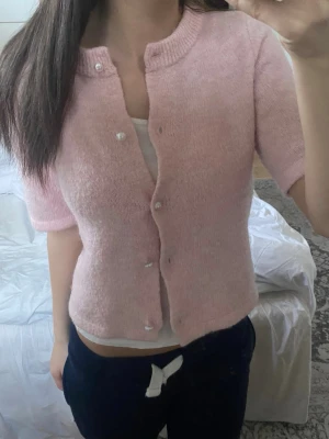 Ljusrosa kofta med pärlknappar - Söt ljusrosa kofta med korta ärmar och rund halsringning. Koftan har pärlknappar framtill och är stickad i ett mjukt material som ger en mysig känsla. Perfekt att slänga över ett linne för en chill look. Därimot är em knapp lite hängande 👌