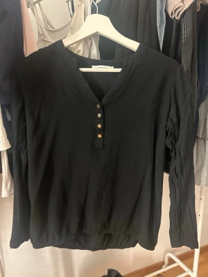 Svart blus med knappar - Stilren svart blus från Selected Femme med v-ringning och fyra dekorativa guldknappar framtill. Blusen har långa ärmar och lätt rynkad nederkant. Tillverkad i ett mjukt, tunt tyg som ger en avslappnad men ändå chic look.