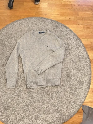 Grå stickad tröja från Polo Ralph Lauren - Snygg grå stickad tröja från Polo Ralph Lauren med rund halsringning och klassisk marinblå logga broderad på bröstet. Tröjan har ribbade muddar vid ärmslut och nederkant!