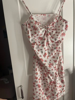 Vit blommig kortklänning med knyt - Söt vit kortklänning med tunna axelband och blommigt mönster i rosa och grönt. Klänningen har en fin knytning framtill vid bysten och är gjord i ett ribbat bomullstyg som känns mjukt och bekvämt. Perfekt för varma dagar och somriga vibes.