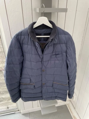 Massimo Dutti vårjacka - Snygg blå pufferjacka med ribbad mörkgrå krage och knäppning framtill. Jackan har två stora fickor med lock och en bröstficka. Quiltad design och diskreta detaljer i brunt vid fickorna. Perfekt för kalla vinterdagar. L men passar även till M