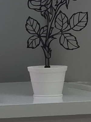 Svart väggdekor i vit kruka - Dekorativ svart ros i plast med stiliserade blad, placerad i en enkel vit plastkruka. Perfekt som prydnad på hylla eller skrivbord. Modern och stilren design som passar i de flesta rum.