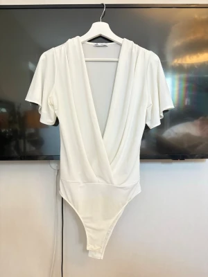 Vit omlottbody från Zara - Snygg vit body från Zara med omlott-skärning och djup V-ringning. Modellen har korta, vida ärmar och mjukt, stretchigt material som sitter bekvämt. Bodyn är vanlig där man knäpper, alltså inte String. Den har en liten liten fläck men går nog bort med lite vanish!