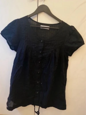 Svart blus med knappar och puffärm - Svart blus med korta puffärmar, rund halsringning och knappar framtill. 