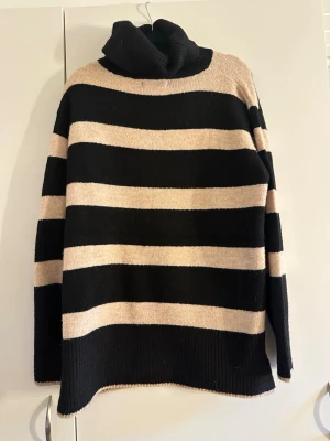 Randig polotröja från Hailys - Stickad polotröja från Hailys i stl S med breda svarta och beige ränder. Tröjan har lång ärm, hög polokrage och avslappnad passform. Perfekt för dig som gillar en klassisk men trendig look.