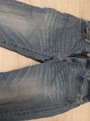 Ljusblå raka jeans med vintage-look - Säljer ett par ljusblå jeans med rak passform och klassisk femficksdesign. Jeansen har en snygg tvättad vintage-look med subtila slitningar och kontrastsömmar. Perfekta för en avslappnad och trendig stil.