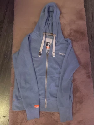 Blå zip hoodie från Superdry - Snygg ljusblå hoodie från Superdry med dragkedja framtill och vita snören i huvan. Broderad Superdry-logga på bröstet och orangea detaljer vid ärmslutet. Mjuk bomullsmix och kängurufickor gör den både chill och praktisk. Perfekt till vardagslooken!