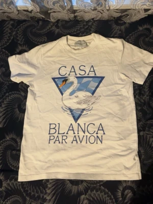 Vit Casa Blanca t-shirt med svan - Vit t-shirt från Casa Blanca med tryck av en svan och blå triangeldetalj på bröstet. Texten 'Casa Blanca Par Avion' i blått framtill. Klassisk passform och rund hals. Perfekt för en stilren och trendig look.