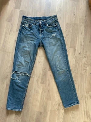 Blå slitna jeans med hål - Ett par blå jeans med raka ben och slitningar, inklusive ett stort hål vid vänster knä och mindre slitningar vid fickorna. Jeansen har klassisk femficksdesign och är gjorda i denim. Perfekta för en avslappnad och trendig look.